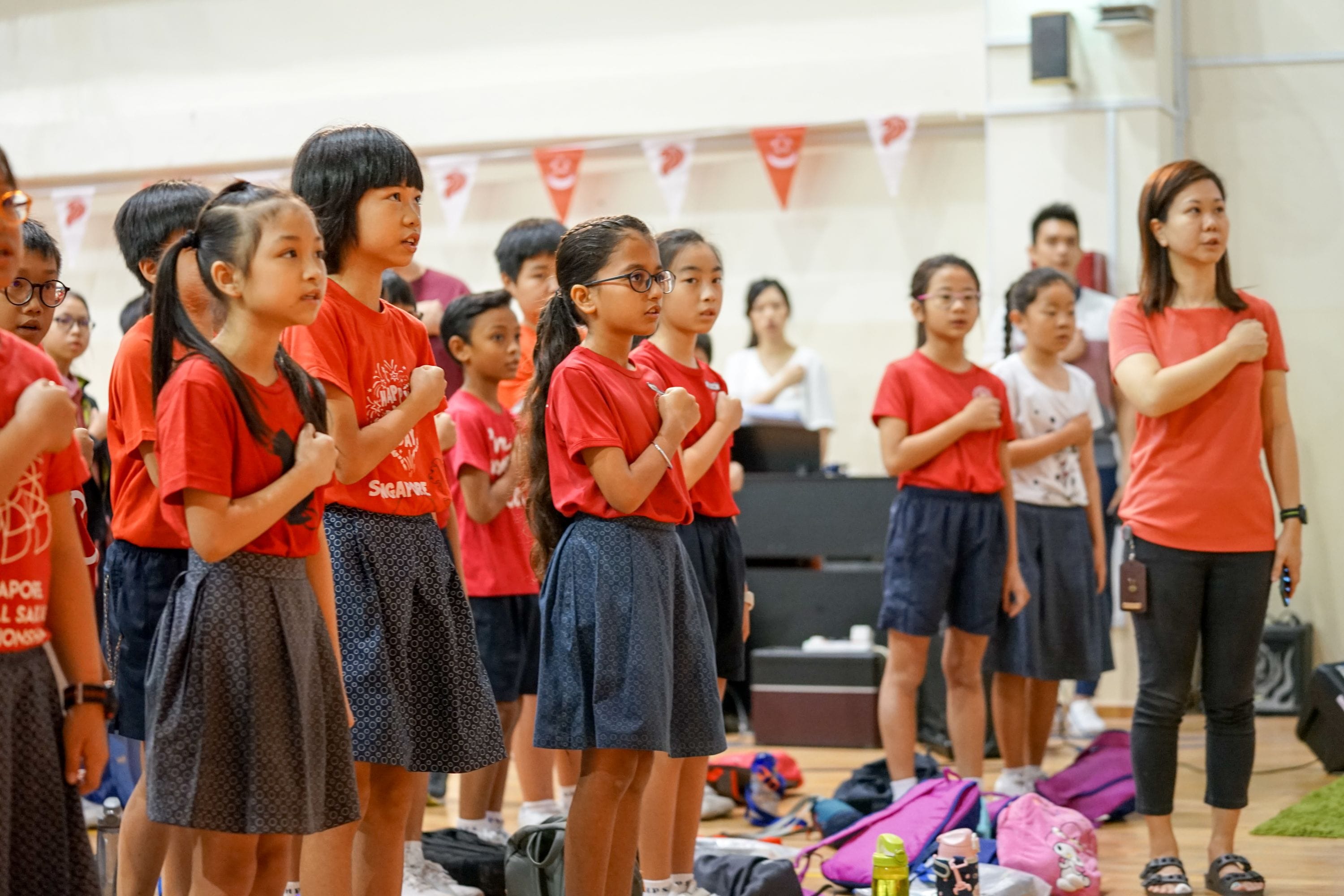 Children and a teacher in red shirts stand with fists on chests in a hall decorated with Singapore flags.